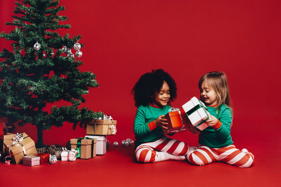 wo children in festive green pajamas and striped pants exchanging Christmas gifts next to a decorated tree, with a red background and wrapped presents around them.