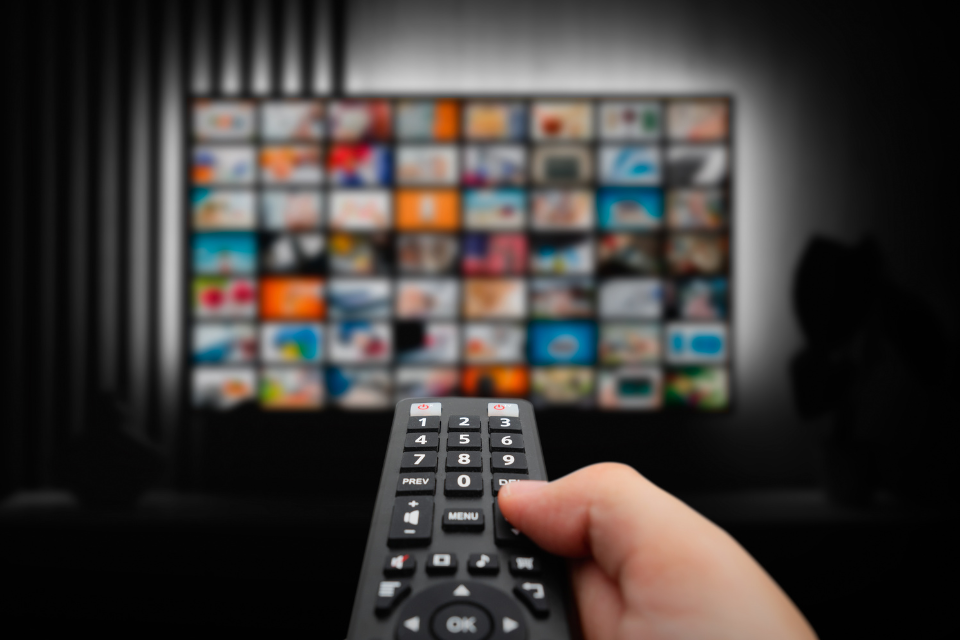 Close-up of a hand holding a TV remote control, pointing towards a blurred screen displaying various streaming service thumbnails in a dark room, symbolizing the concept of streaming entertainment.